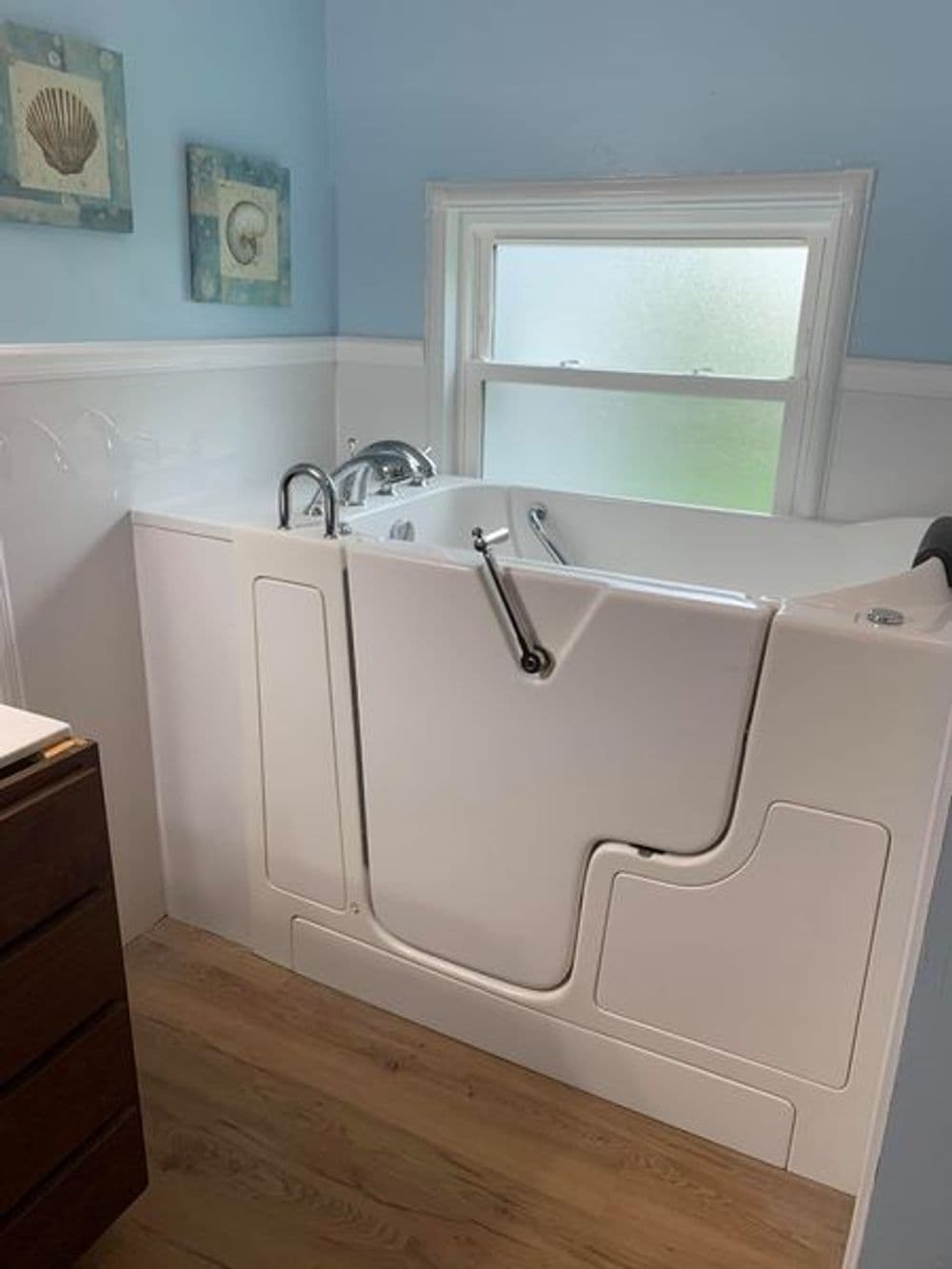 Walk-in bathtub in a bright bathroom with light blue walls and wooden flooring.