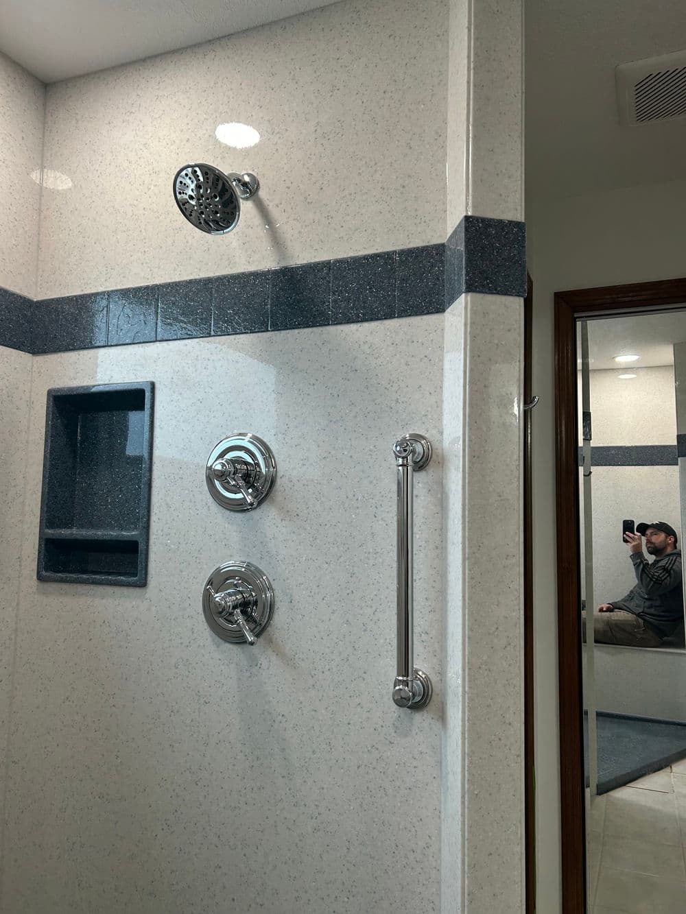 Modern shower with chrome fixtures, grab bar, and glass tile accent wall reflecting a person in the mirror.